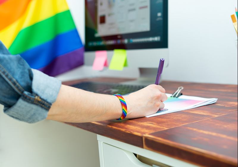 A close-up of a woman's hand. She is writing on a piece of paper. She is using a purple pen. On her wrist is a rainbow bracelet. There is a Pride flag in the background.
